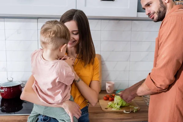 Hombre cocina ensalada cerca sonriente esposa e hijo en la cocina - foto de stock