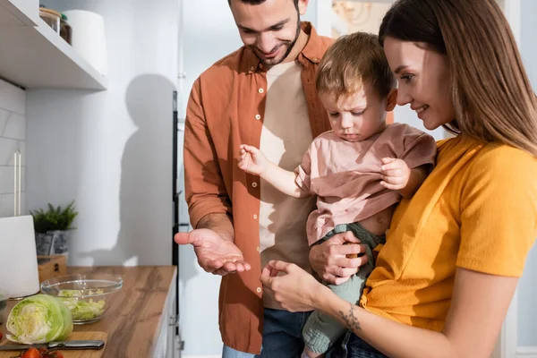 Щасливі батьки дивляться на дитину біля овочів на кухонній стільниці — стокове фото