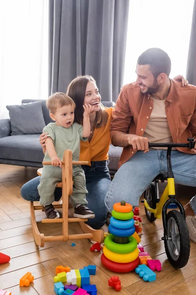 Mulher alegre abraçando filho no cavalo de balanço e marido de bicicleta em casa — Fotografia de Stock