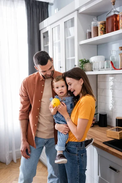 Padres felices mirando a su hijo pequeño con limón fresco en la cocina - foto de stock