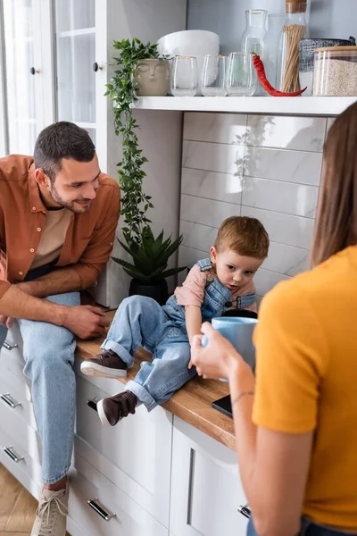 Mujer borrosa sosteniendo taza cerca de marido e hijo en la cocina - foto de stock