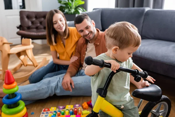 Niño de pie cerca de la bicicleta y los padres borrosos en la sala de estar - foto de stock