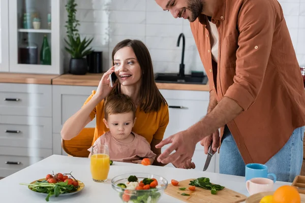 Young woman talking on smartphone near son and husband cooking salad at home — Stock Photo