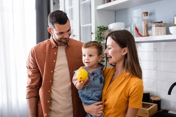 Positive parents looking at toddler son with lemon in kitchen — Stock Photo