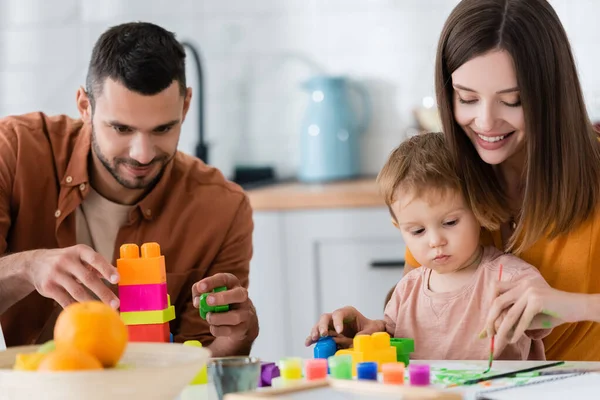 Mujer sonriente acercándose hijo y marido con bloques de construcción en casa - foto de stock