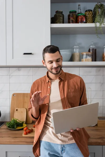 Sorrindo homem ter chamada de vídeo no laptop perto de legumes na cozinha — Fotografia de Stock