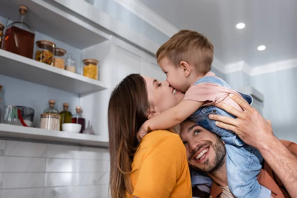 Sonriente mujer besando hijo cerca de marido en casa - foto de stock