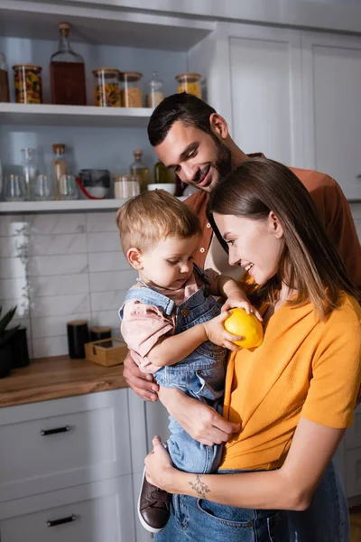 Padres jóvenes mirando a un niño pequeño con limón en la cocina - foto de stock