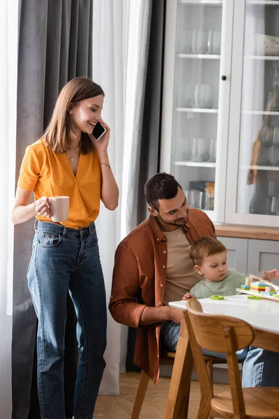 Frau mit Tasse telefoniert auf Smartphone, während Mann und Sohn zu Hause spielen — Stockfoto