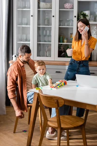 Mujer hablando en el teléfono inteligente y sosteniendo la taza cerca de la computadora portátil y la familia jugando juego en casa - foto de stock