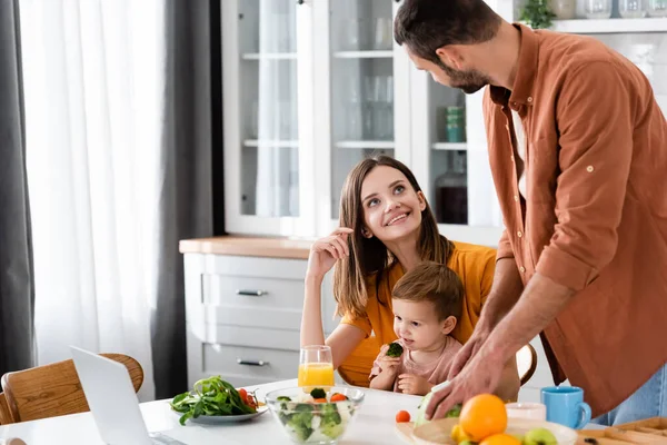 Улыбающаяся женщина смотрит на мужа, готовящего салат рядом с ребенком и ноутбук на кухне — стоковое фото