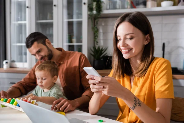 Freelancer positivo usando smartphone perto de laptop e jogo de família em casa — Fotografia de Stock