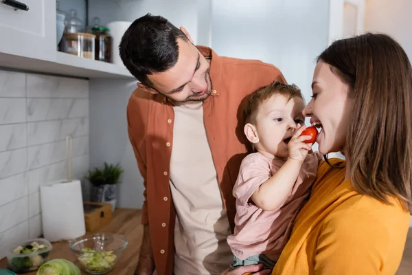 Cheerful man standing near son feeding mother with cherry tomato in kitchen — Stock Photo