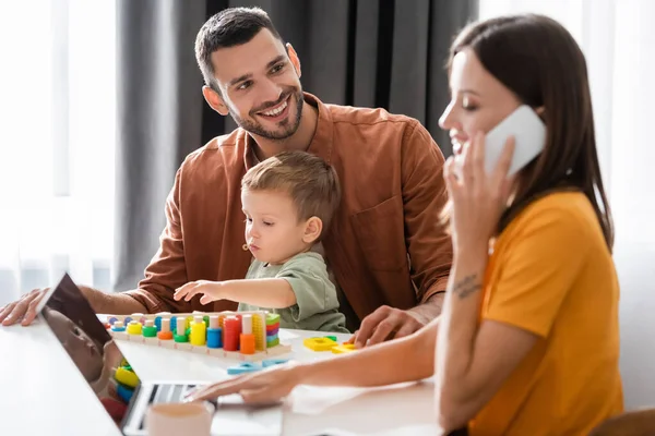 Sorrindo homem olhando para esposa desfocada falando no smartphone e usando laptop perto do filho e jogo em casa — Fotografia de Stock