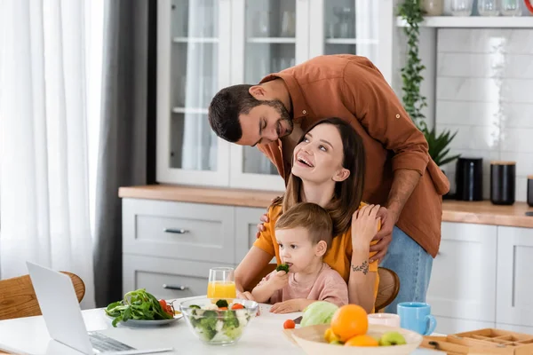 Un homme positif étreignant sa femme près de son fils, sa salade et son ordinateur portable dans la cuisine — Photo de stock