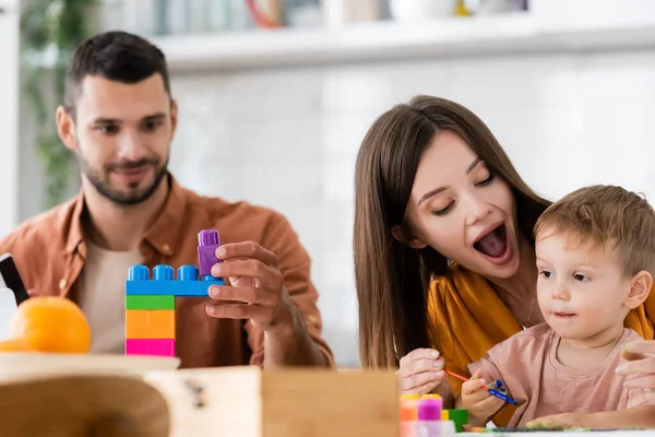 Mulher segurando pincel perto do filho e do marido com blocos de construção em casa — Fotografia de Stock