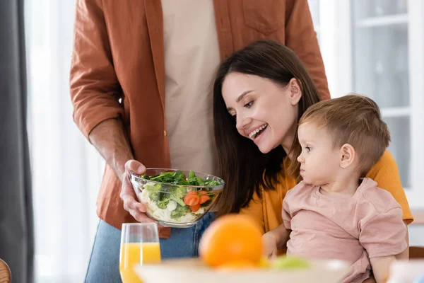 Hombre sosteniendo ensalada fresca cerca alegre esposa e hijo en casa - foto de stock