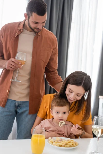 Homem segurando vinho perto da esposa alimentando filho com massa em casa — Fotografia de Stock