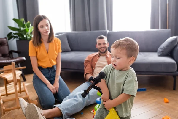 Toddler child holding bike near blurred parents and rocking horse at home — Stock Photo