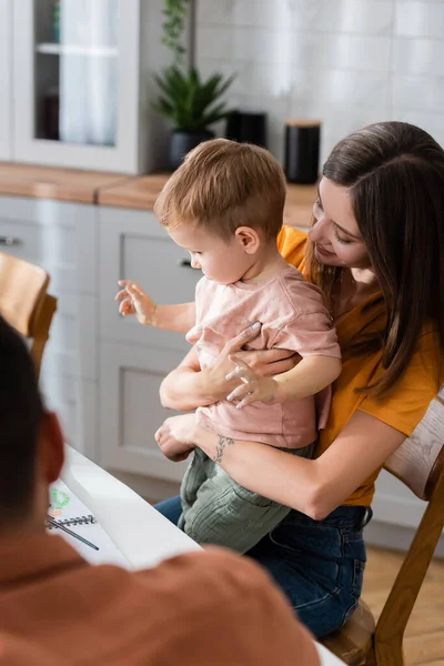 Lächelnde Frau umarmt Sohn neben Skizzenbuch und verschwommenem Ehemann zu Hause — Stockfoto