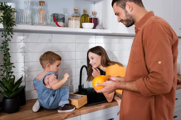 Sorrindo pais segurando laranjas perto do filho na bancada da cozinha — Fotografia de Stock