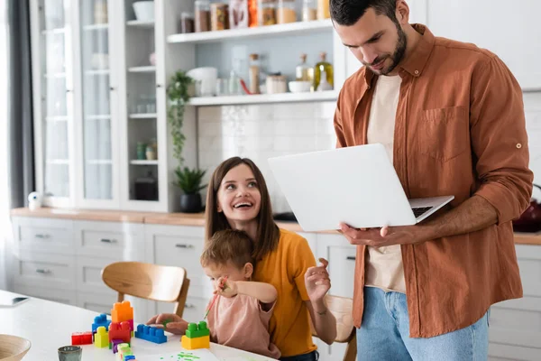 Hombre usando portátil cerca sonriente esposa e hijo con bloques de construcción en casa - foto de stock