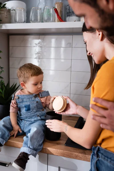 Padres sosteniendo la tapa y el frasco cerca del niño pequeño en la cocina - foto de stock