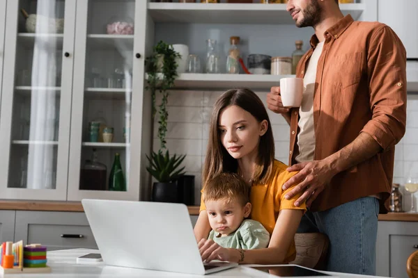 Hombre sosteniendo la taza cerca de la esposa usando el ordenador portátil y el niño en la cocina - foto de stock