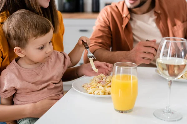 Niño pequeño sosteniendo tenedor cerca de los padres y pasta en la cocina - foto de stock