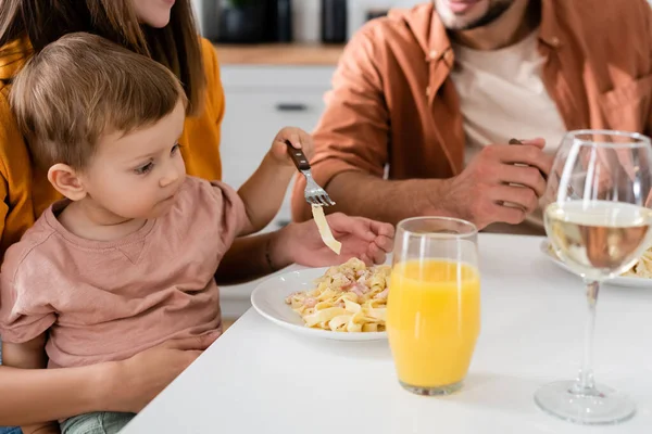 Enfant en bas âge tenant fourchette près des parents et des pâtes dans la cuisine — Photo de stock