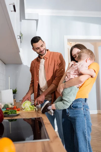 Femme tenant tout-petit garçon près de mari coupe salade dans la cuisine — Photo de stock