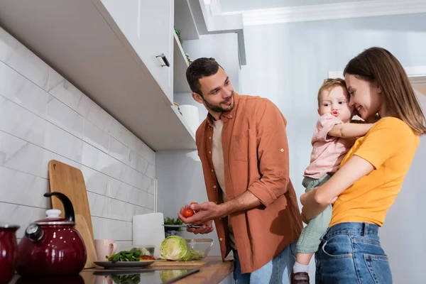 Madre positiva abrazando hijo mientras marido cocina ensalada en la cocina - foto de stock