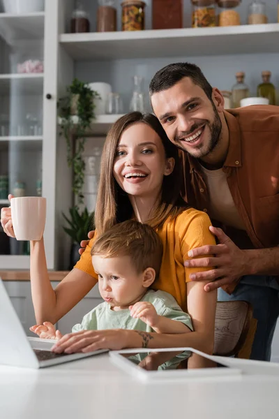 Pais alegres olhando para a câmera perto de dispositivos e filho em casa — Fotografia de Stock