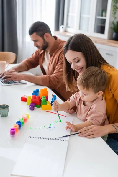 Sourire femme peinture avec fils près de blocs de construction et mari en utilisant ordinateur portable à la maison — Photo de stock