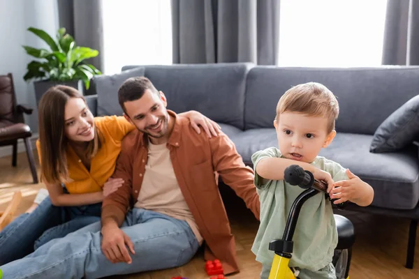 Niño de pie cerca de la bicicleta y los padres borrosos en la sala de estar - foto de stock