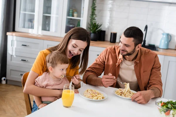 Donna sorridente che alimenta la pasta a figlio vicino a marito e succo d'arancia in cucina — Foto stock