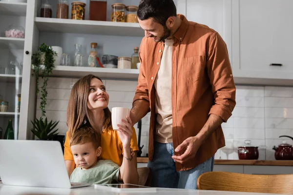 Mujer sonriente sosteniendo taza cerca de marido, hijo y dispositivos en casa - foto de stock