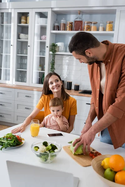 Hombre cortando repollo cerca sonriente esposa e hijo en cocina - foto de stock