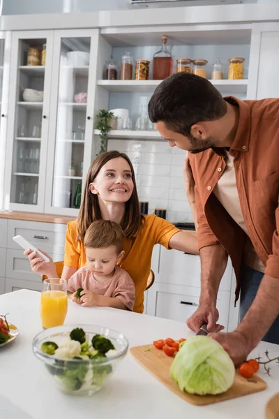 Smiling woman holding smartphone near husband cooking salad and son at home — Stock Photo