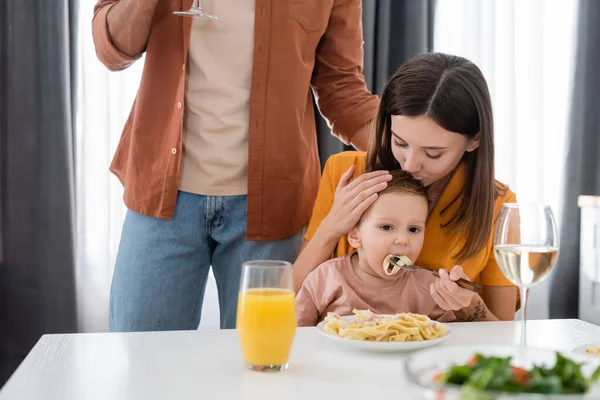 Женщина целует и кормит ребенка возле макарон и мужа дома — стоковое фото