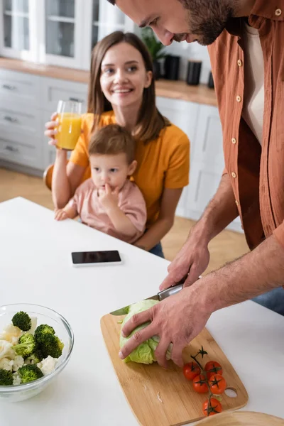 Young man cooking salad near family and smartphone on table at home — Stock Photo