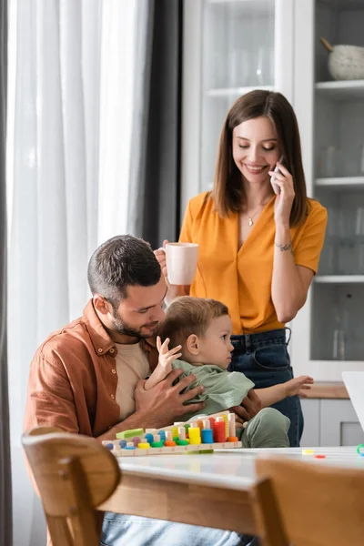 Padre giocare con figlio vicino gioco educativo e moglie parlando su smartphone a casa — Foto stock