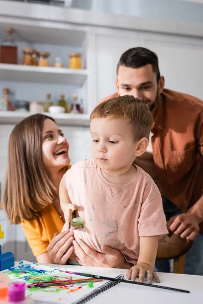 Niño pequeño cerca de cuaderno de bocetos, pinceles y padres en casa - foto de stock