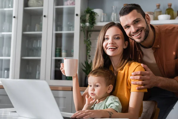 Famiglia sorridente guardando la fotocamera vicino al figlio e laptop a casa — Foto stock