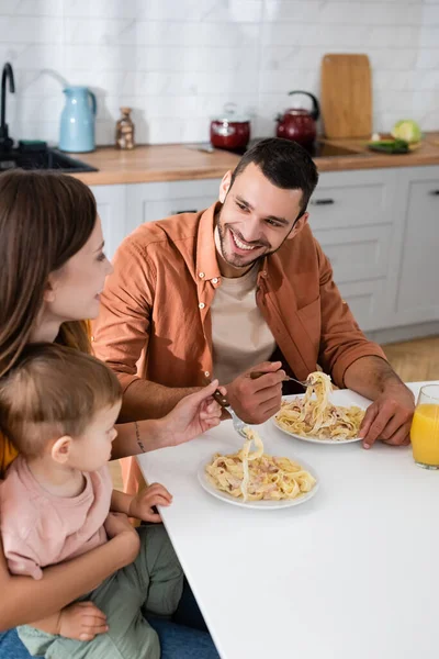 Hombre sonriente mirando a esposa e hijo cerca de pasta y jugo de naranja en casa - foto de stock