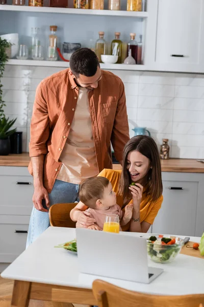 Famiglia positiva guardando bambino bambino vicino laptop e insalata in cucina — Foto stock