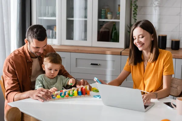 Freelancer en utilisant un ordinateur portable près de mari et fils jouer à un jeu éducatif — Photo de stock