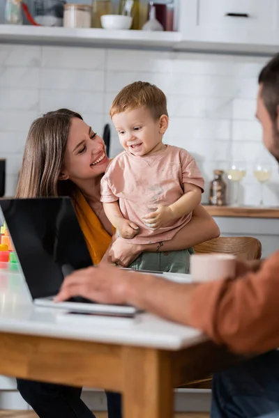 Feliz mãe abraçando criança enquanto marido usando laptop em casa — Fotografia de Stock