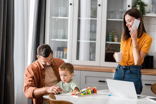 Smiling father playing with child near wife with cup talking on smartphone — Stock Photo