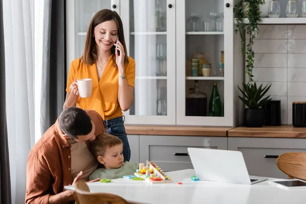 Улыбающаяся женщина разговаривает по смартфону рядом с мужем, играет с сыном на кухне — стоковое фото